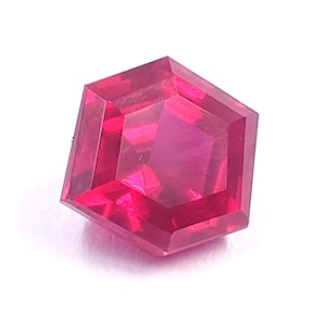 May include: A vibrant, transparent, hexagonal-cut gemstone with a deep magenta-red hue. The stone features a faceted surface that reflects light, creating internal patterns and depth within its six-sided geometric shape and polished edges.