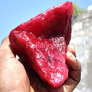 Puede incluir: Una gran gema de rubí en bruto, de color rojo intenso. La piedra en bruto tiene una superficie texturizada y se sostiene en una mano. El texto "Rohangems" es visible en la piedra.