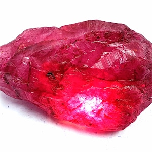 Könnte beinhalten: Ein großer, tief roter Rubin-Kristall mit einer rauen, facettierten Oberfläche. Der Kristall wird von unten beleuchtet, wodurch seine innere Struktur hervorgehoben wird.