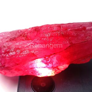 May include: A raw ruby gemstone, displaying a deep red colour with lighter pink areas. The uncut mineral has a crystalline structure and a natural, unpolished surface. The image shows the gemstone's natural beauty. The gemstone is a deep, saturated red colour.