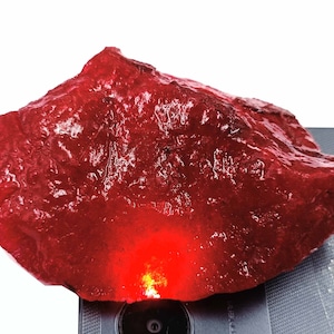 May include: A large, rough, red gemstone with a deep, rich color. The gemstone is illuminated from below, highlighting its internal structure.
