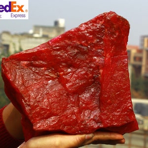 May include: A large, rough-hewn, red gemstone with a textured surface. The stone is held in a person's hands, with a blurred background of buildings and sky. The FedEx Express logo is visible in the upper left corner.