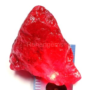 May include: A raw, uncut ruby gemstone with a vibrant red hue. The rough surface of the gem is visible, and the image includes the text "Rohangems". The gemstone is placed on a measuring tool.