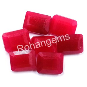 Pode incluir: Seis cabochões de pedras preciosas vermelhas em corte retangular esmeralda. As pedras preciosas são lisas e polidas, com um leve brilho. As pedras preciosas estão dispostas em uma pilha solta.