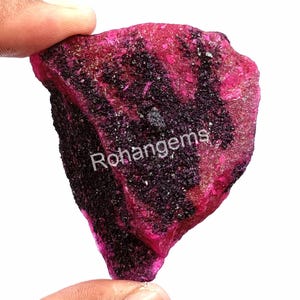Puede incluir: Una gran gema de rubí rosa en bruto con inclusiones negras. La gema se sostiene en una mano, y la palabra "Rohangems" es visible en la imagen.
