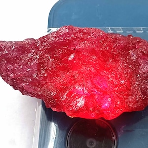 May include: A large, rough, red ruby gemstone. The ruby is a deep red color and has a rough, natural surface.