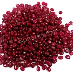Puede incluir: Un montón de piedras preciosas de rubí pulidas, de forma ovalada. Las gemas son de un color rojo intenso y rico y varían ligeramente de tamaño. Las piedras preciosas están dispersas sobre una superficie blanca.