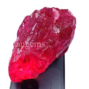 Peut inclure: Une grosse pierre précieuse de rubis brute, d'un rouge profond. La surface de la pierre est texturée et réfléchit la lumière, avec le texte "tangems" visible. La pierre est posée sur un fond sombre.