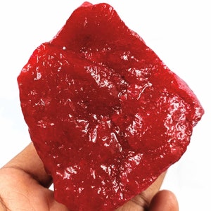 May include: A rough-cut, vibrant red gemstone with a glossy, reflective surface. The irregularly shaped stone has a natural, organic form, showcasing its raw, unpolished state. The gemstone is held in a hand against a white background.