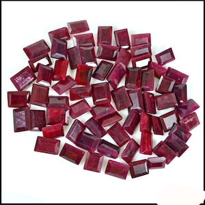 Puede incluir: Una colección de piedras preciosas de rubí rojo facetadas y sueltas, de forma rectangular. Los rubíes están esparcidos sobre una superficie blanca.