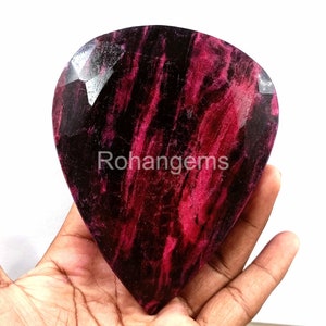 May include: A large, heart-shaped, faceted ruby gemstone with a deep red and black colour. The gemstone has a striated pattern and is held in a hand.
