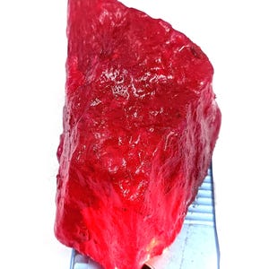May include: A raw, uncut ruby gemstone with a deep red color. The rough stone has a textured surface and is positioned next to a ruler for scale. This natural gemstone is ideal for jewelry making or collecting.