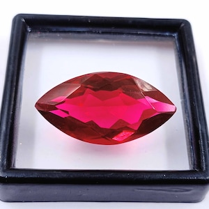 May include: A vibrant, deep red marquise-shaped gemstone with a faceted cut, displayed in a black square frame.