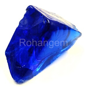 May include: A large, irregularly shaped piece of deep blue glass. The surface has a glossy sheen, with internal swirls and variations in color. The word "Rohangem" is printed on the surface. The glass appears to be a decorative or collectible item.