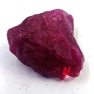 Pode incluir: Uma gema de rubi bruta, não lapidada, com um tom vermelho-púrpura profundo. A pedra natural tem uma superfície texturizada com estruturas cristalinas visíveis. Uma pequena área deixa passar a luz, revelando sua cor vibrante. O rubi está sobre uma superfície branca.