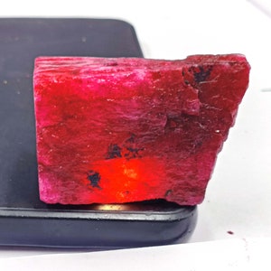 May include: A raw, vibrant red ruby gemstone specimen with a rough, textured surface and visible dark inclusions. The stone displays a deep crimson hue with a bright, concentrated orange-red glow in the center, resting on a black rectangular object.