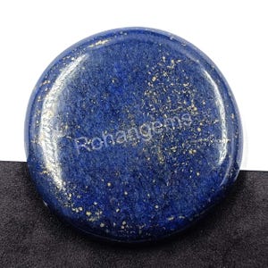 Pode incluir: Uma pedra preciosa redonda de lápis-lazúli polida, de cor azul profunda com manchas douradas. O texto "Rohangems" está impresso na superfície da pedra.