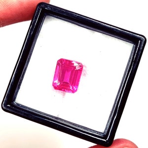 May include: A vibrant, rectangular-cut, hot pink gemstone is displayed within a black framed case with a white interior. The gemstone is the focal point, showcasing its facets and color. The case is held by a person's fingers.