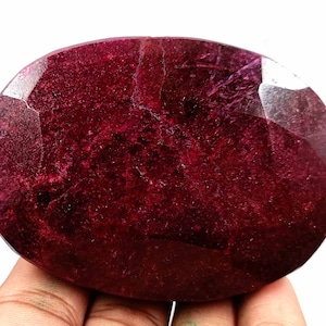 May include: A large, oval-shaped, deep red ruby gemstone with a faceted cut. The gemstone has a slightly cloudy appearance.
