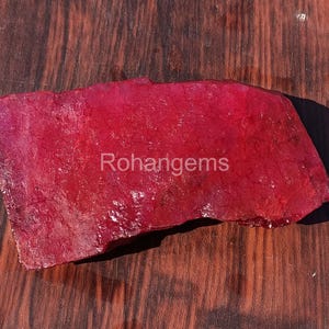 以下が含まれることがあります： 「Rohangems」の白文字が入った、ざらざらとした深紅色の宝石。石は質感のある表面を持ち、暗い木製の背景に置かれています。宝石の色は鮮やかで鮮やかな赤色です。