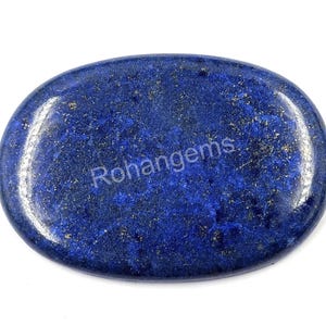 Pode incluir: Pedra preciosa oval em lápis-lazúli com superfície polida. A pedra azul escura tem manchas douradas e o texto "Rohangems" é visível. A pedra preciosa está sobre um fundo branco.