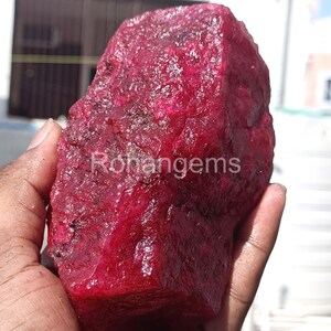 Puede incluir: Un gran rubí en bruto, de color rojo intenso, sostenido en una mano. La gema tiene una superficie texturizada. El texto "Rohangems" es visible en la superficie. La imagen está tomada con luz natural.