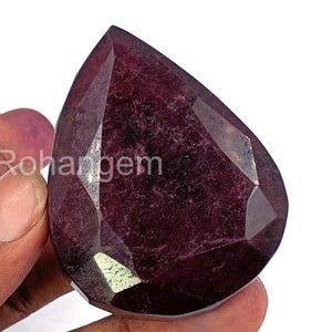 May include: A faceted, pear-shaped, deep red gemstone. The gem has a polished surface with multiple facets, reflecting light. The text "Rohangem" is visible on the left side of the gem. The gem is held by a person's hand.