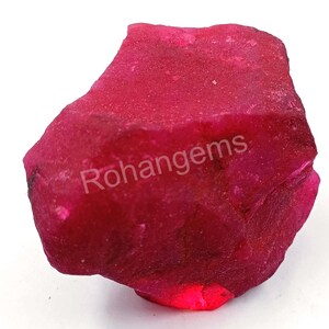 Può includere: Un grande rubino grezzo di colore rosso. Il rubino è di un rosso intenso e ricco e ha una forma leggermente irregolare. La pietra preziosa non è lucidata e ha un aspetto naturale e grezzo. Rohangems è stampato sul rubino.