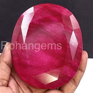 Puede incluir: Una gran gema de rubí facetada con un tono rojo intenso. La piedra de forma ovalada tiene múltiples facetas que reflejan la luz, creando un efecto brillante. El texto "Rohangems" es visible.