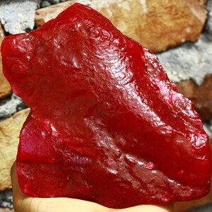 May include: A large, rough, red ruby gemstone. The ruby is a deep, rich red color and has a glossy finish.