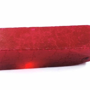 Puede incluir: Una gema de rubí en bruto, de forma rectangular y color rojo intenso. La superficie es rugosa y texturizada, con grietas e imperfecciones visibles. El rubí se presenta sobre un fondo blanco liso.