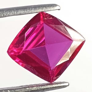 May include: A vibrant, cushion-cut gemstone with a deep, saturated magenta-pink color. The stone features a faceted surface that reflects light, creating distinct geometric patterns across its top. It is held securely by metal tweezers.