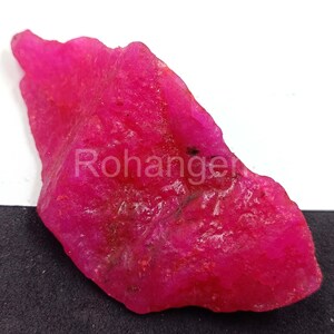 Puede incluir: Una gema de rubí rosa vibrante y en bruto con una superficie texturizada. El rubí tiene una forma irregular y se muestra sobre un fondo blanco y negro. La palabra "Rohanger" está impresa en la gema.