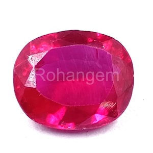 Puede incluir: Una gema de rubí ovalada y facetada. La gema es de un rojo intenso y vibrante con un interior rojo rosado más claro. El texto "Rohangem" está impreso en la superficie de la gema.
