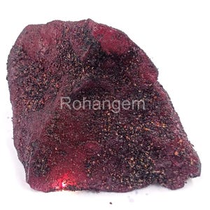 May include: A rough-textured, dark red gemstone with a dusting of black and tan particles. The word "Rohangem" is printed on the surface. The stone has an irregular shape and is set against a white background.