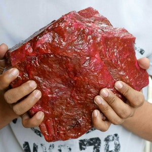May include: A large, rough, red ruby gemstone. The ruby is being held in the hands of a person.