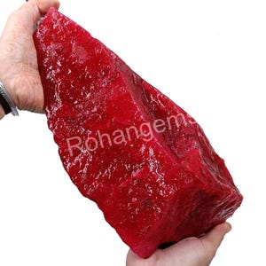 May include: A large, rough, red ruby gemstone. The ruby is in the shape of a large, irregular rock.