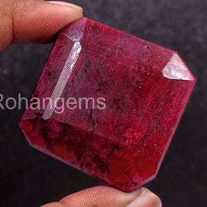 May include: A faceted, rectangular ruby gemstone with a deep red hue. The gem is held up by a hand, and the text "Rohangems" is visible on the surface. The gemstone has a polished, reflective surface.