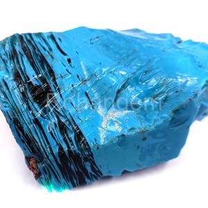 May include: A vibrant, irregularly shaped turquoise gemstone with streaks of black. The polished surface reflects light, showcasing the stone's natural texture and color variations. The gemstone is set against a white background.
