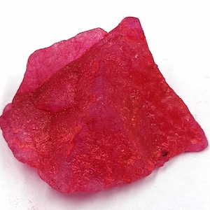 Op de afbeelding: Een grote, ruwe, roze robijnkristal met een diepe rode tint. De kristal heeft een natuurlijke, ongepolijste uitstraling.