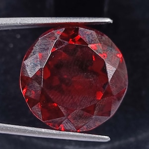 Puede incluir: Una piedra preciosa de color rojo intenso con un corte facetado. La piedra preciosa es redonda y tiene un alto pulido.