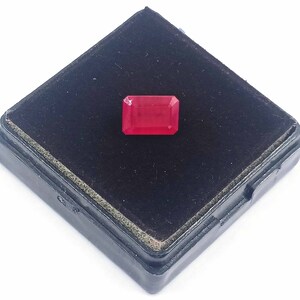 Puede incluir: Una piedra preciosa de color rojo intenso con forma rectangular, colocada en una caja de terciopelo negro.