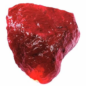 Può includere: Una gemma di rubino grezza, non tagliata, di colore rosso intenso. La superficie della pietra ha un aspetto ruvido e strutturato, con una leggera lucentezza. Il rubino è posto su uno sfondo bianco, evidenziando la sua forma naturale.