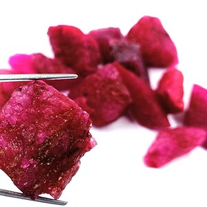 May include: A close-up of a large, deep red ruby gemstone held by tweezers. The ruby is rough and unpolished, with a crystalline structure. Several smaller, similarly colored rubies are scattered in the background.