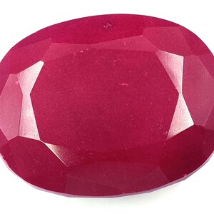 May include: A faceted, oval-shaped ruby gemstone. The gem is a deep, saturated red colour with multiple facets that reflect light. The gemstone is set against a white background.
