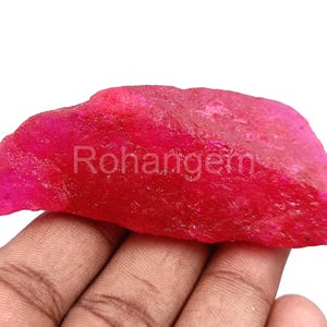 May include: A raw, uncut ruby gemstone in a vibrant pinkish-red colour. The surface has a textured appearance. The word "Rohangem" is printed on the surface of the gemstone.