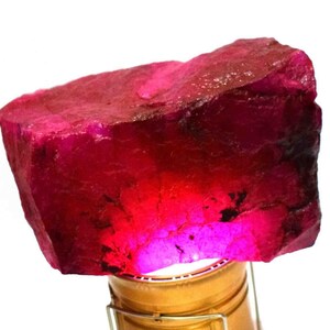 Puede incluir: Una gran gema de rubí en bruto, de color rojo intenso. El rubí está iluminado desde abajo, mostrando su textura natural y variaciones de color. La gema se exhibe sobre un soporte dorado sobre un fondo blanco.