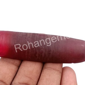 May include: A raw ruby gemstone, elongated with a deep red colour at one end, fading to a darker, purplish-red. The surface has a natural, unpolished texture. The text "Rohangems" is visible on the surface.