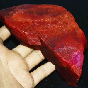 May include: A large, rough, red ruby gemstone. The ruby is a deep red colour with some areas of a lighter pink hue. The ruby is in the shape of a crescent moon.
