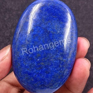Pode incluir: Uma pedra preciosa de lápis-lazúli polida, em forma oval. A pedra azul escura tem manchas douradas e um acabamento brilhante. O texto "Rohangems" é visível. A pedra preciosa é segurada em uma mão.